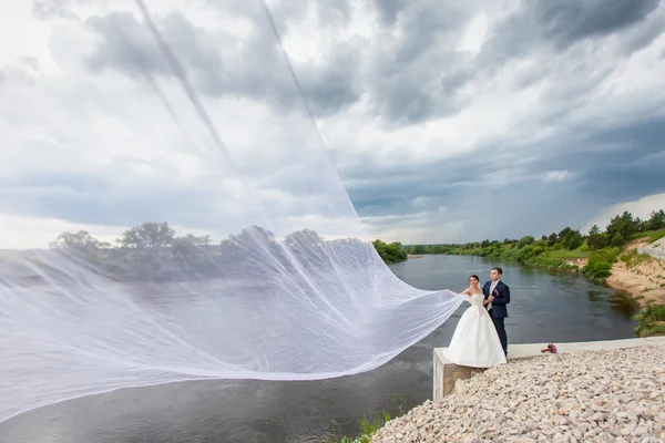 Marié et mariée avec un très long voile nuptial debout sur la rive de la rivière — Photo