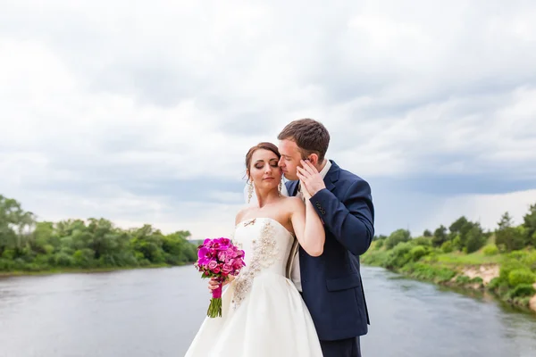 O noivo está beijando a noiva na margem do rio — Fotografia de Stock