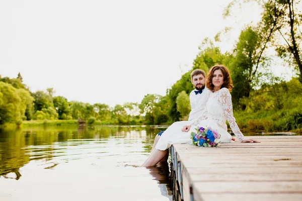 Braut und Bräutigam sitzen auf einem hölzernen Steg am Teich — Stockfoto