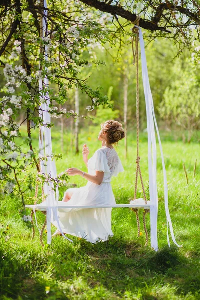 Jonge bruid met blond haar in witte negligé met behulp van parfum op een touw swing — Stockfoto