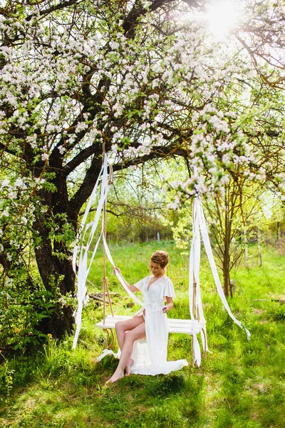 Jonge bruid met blond haar in wit negligé zitten op een touw swing — Stockfoto