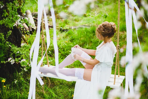 Giovane sposa con i capelli biondi in negligee bianco e calze mettere su una giarrettiera — Foto Stock