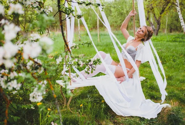 Jonge bruid met blond haar in wit negligé en kousen zitten op een touw swing — Stockfoto