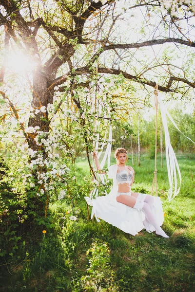 Giovane sposa con i capelli biondi in negligee bianco e calze in posa su un'altalena corda — Foto Stock