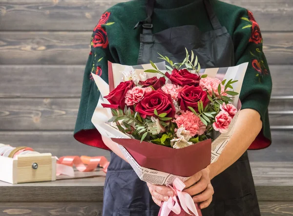 In the hands of the florist, a bouquet of red roses, eucalyptus and carnations. A bouquet in a wrapper. Florist in an apron, bouquet at arm\'s length. Florist in a flower shop. Background wooden table and wood panel on the wall