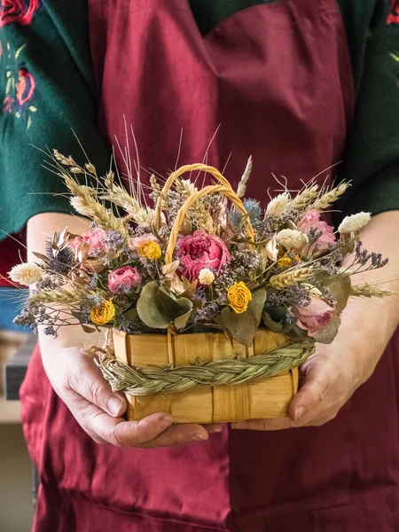 In the hands of the florist, a bouquet of dried flowers with a rose, wheat, eucalyptus in a wicker basket with handles, decorated with a scythe of grass. Background apron at the florist fuchsia color