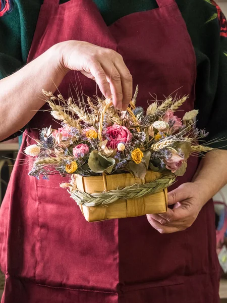 In the hands of the florist, a bouquet of dried flowers with a rose, wheat, eucalyptus in a wicker basket with handles, decorated with a scythe of grass. Background apron at the florist fuchsia color