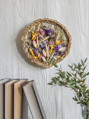 Duvarda el yapımı bir kurutulmuş çiçek, çiçek, buğday, ot ve örgü paneli asılı. Çerçevenin altında kitaplar, vazo dolusu su ve yeşil yapraklarla dolu bir raf var. Hafif ahşap bir arka planda.