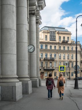Şehir sokağı. Gri kolonları ve antika saati olan bir bina. İnsanlar, yol işaretleri, asfalt yol. Güzel mimari. Arka planda sarı ev.