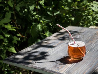 Bahçedeki koyu ahşap bir masada, bardakta elma şarabı. Arka planda yeşil çalılar ve ağaçlar var. Güneş ışığı. Cama bir göz at. Masadaki ağaçların yapraklarının gölgesi..