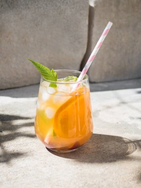 Ahududu yapraklı soğuk portakal kokteyli, buz. Bardakta kokteyl ve pipet. Arka plan beton kaplı doğal bir beton bloktur. Güneş ışığı, ağaç yapraklarının gölgesi