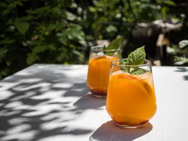 Bahçedeki beyaz ahşap bir masada, ahududu yapraklı ve buzlu soğuk turuncu bir kokteyl. Arka plan - bahçedeki ağaçların yaprakları. Güneş ışığı, masadaki ağaçların yapraklarının gölgesi..