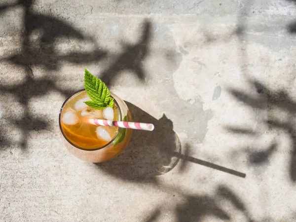 Cold orange cocktail with raspberry leaves, ice. Cocktail in glass with cocktail straw. View from above. The background is a natural concrete block with cement. Sunshine, shadow from tree foliage