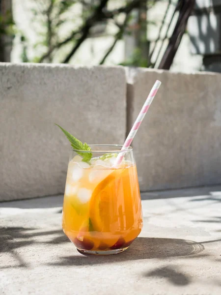 Cold orange cocktail with raspberry leaves, ice. Cocktail in glass with cocktail straw.  The background is a natural concrete block with cement. Sunshine, shadow from tree foliage
