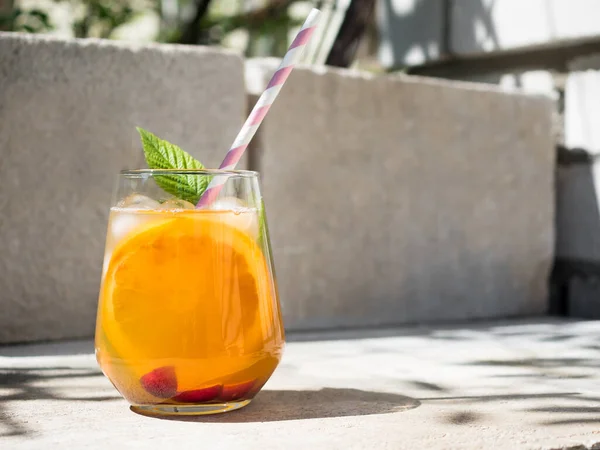Cold orange cocktail with raspberry leaves, ice. Cocktail in glass with cocktail straw.  The background is a natural concrete block with cement. Sunshine, shadow from tree foliage