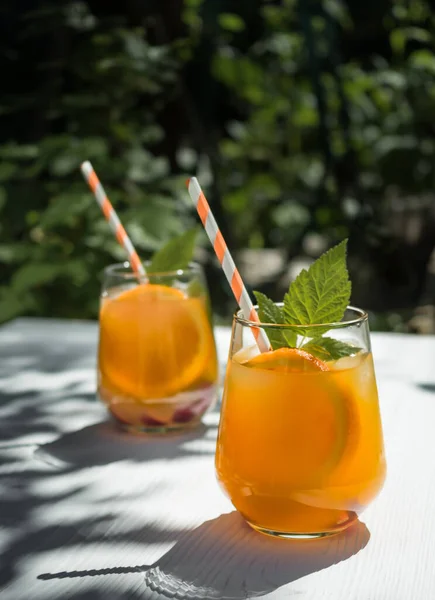 On a white wooden table in the garden, a cold orange cocktail with raspberry leaves and ice. Orange cocktail tubes. Background - foliage from trees in the garden. Sunlight, shadow from the foliage of trees on the table.