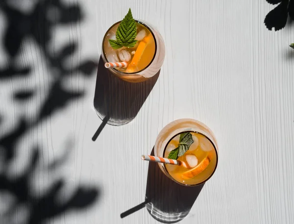 Cold orange cocktail with raspberry leaves, ice. Cocktail in a glass with a cocktail tube. View from above. Background - a white wooden table. Sunshine, shadow from tree foliage