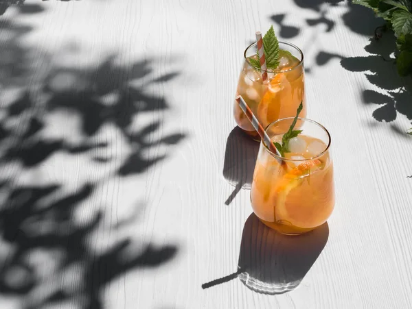 Cold orange cocktail with raspberry leaves, ice. Cocktail in a glass with a cocktail tube. Background - a white wooden table. Sunshine, shadow from tree foliage