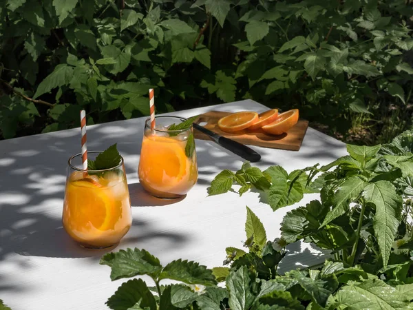 On a white wooden table in the garden, a cold orange cocktail with raspberry leaves and ice. Making cocktails. Background - foliage from trees in the garden. Foliage frame.