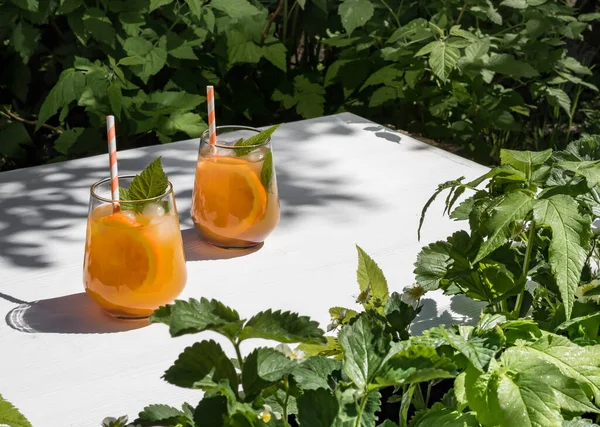 On a white wooden table in the garden, a cold orange cocktail with raspberry leaves and ice. Orange cocktail tubes. Background - foliage from trees in the garden. Foliage frame. Sunlight, shadow from the foliage of trees on the table.