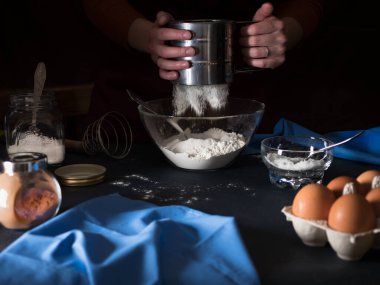 Pasta pişiriyorum. Unu bir kaseye yerleştiriyorum. Masanın üzerinde turta yapmak için malzemeler (un, tereyağı, şeker, tuz, kakao, yumurta, maya), mutfak gereçleri ve tekstil. Arka planda önlüklü bir kız var. Koyu arkaplan