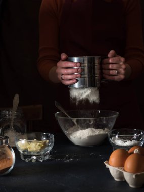 Pasta pişiriyorum. Unu bir kaseye yerleştiriyorum. Masanın üzerinde turta yapmak için malzemeler vardır (un, tereyağı, şeker, tuz, kakao, yumurta, maya), mutfak gereçleri. Arka planda önlüklü bir kız var. Koyu arkaplan