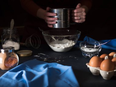 Pasta pişiriyorum. Unu bir kaseye yerleştiriyorum. Masanın üzerinde turta yapmak için malzemeler (un, tereyağı, şeker, tuz, kakao, yumurta, maya), mutfak gereçleri ve tekstil. Arka planda önlüklü bir kız var. Koyu arkaplan
