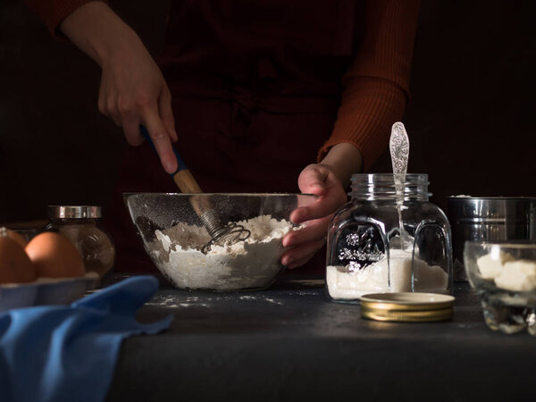 Cooking a cake. On the table are ingredients for making a pie (flour, eggs, cocoa, butter, sugar, salt), kitchen utensils and textiles. Kneading the dough with a whisk. In the background is a girl in an apron. Dark background