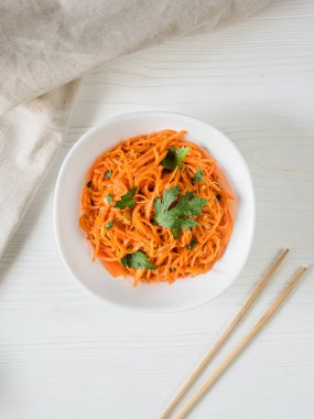 Baharatlı Asya salatası, rendelenmiş havuç ve maydanoz. Beyaz arka planda Kore havuçları ve yemek çubukları ve tekstil dolu beyaz bir kasede. Vejetaryen diyeti. Sağlıklı gıda konsepti. Yukarıdan bak. Düz yatıyordu. Dikey