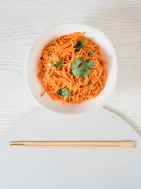 Baharatlı Asya salatası, rendelenmiş havuç ve maydanoz. Beyaz arka planda, çubuklu beyaz bir kâse içinde Kore havuçları. Tabakta salata var. Vejetaryen diyeti. Sağlıklı gıda konsepti. Yukarıdan bak. Düz yatıyordu. Dikey. 