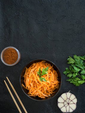 Baharatlı Asya salatası ve rendelenmiş havuç. Siyah arka planda, yemek için yemek çubukları, salata hazırlamak için malzemeler olan koyu bir kasede Kore havuçları. Vejetaryen diyeti. Sağlıklı gıda konsepti. Yukarıdan bak. Uzayı kopyala Dikey. Düz yatıyordu