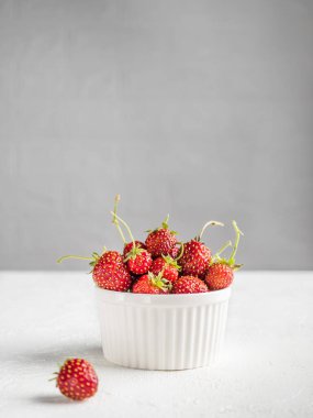 İştah açıcı taze çilekler, kesikler ve dallarla beyaz arka planda beyaz bir kasede yatıyor. Arka planda madalyalı tuğla. Vejetaryen atıştırması. Dikey. Boşluğu Kopyala