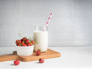 İştah açıcı yeşil pirzolalı taze çilekler beyaz bir kasede ve tahta bir tahtanın üzerinde pipetle bir bardak sütte yatıyor. Arka planda madalyalı tuğla. Kahvaltı konsepti. Yatay olarak. Boşluğu kopyala.