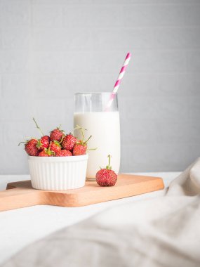 Tahtanın üzerinde, bir bardak sütlü pipet ve kırmızı çilekli beyaz bir kase. Tekstil ön planda. Kahvaltı konsepti. Vejetaryen atıştırmalıkları. Dikey