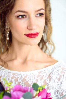 Beyaz örgü elbiseli neşeli bir gelin stüdyoda beyaz bir arka planda bir buket çiçekle poz veriyor.