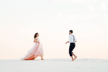 Genç çift, siyah pantolonlu bir adam ve pembe elbiseli bir kız çölün beyaz kumu boyunca yürüyorlar.