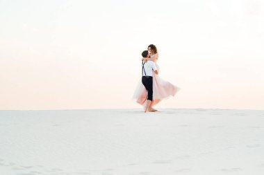 Genç çift, siyah pantolonlu bir adam ve pembe elbiseli bir kız çölün beyaz kumu boyunca yürüyorlar.