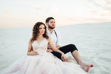 Genç çift, siyah pantolonlu bir adam ve pembe elbiseli bir kız çölün beyaz kumu boyunca yürüyorlar.