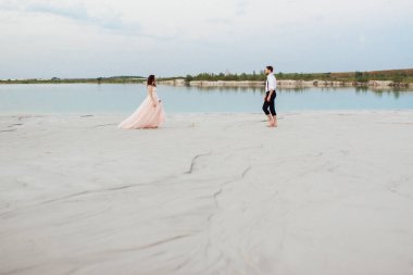 Genç çift, siyah pantolonlu bir adam ve pembe elbiseli bir kız çölün beyaz kumu boyunca yürüyorlar.