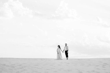 Genç çift, siyah pantolonlu bir adam ve pembe elbiseli bir kız çölün beyaz kumu boyunca yürüyorlar.