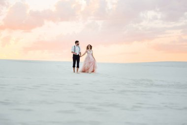 Genç çift, siyah pantolonlu bir adam ve pembe elbiseli bir kız çölün beyaz kumu boyunca yürüyorlar.