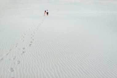 Genç çift, siyah pantolonlu bir adam ve pembe elbiseli bir kız çölün beyaz kumu boyunca yürüyorlar.