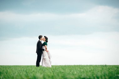 Kahverengi takım elbiseli damat ve fildişi rengi elbiseli gelin yeşil bir tarlada gökyüzüne doğru geri çekiliyor.