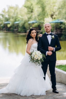 Beyaz elbiseli siyah saçlı bir gelin ve düğün günlerinde parkta papyonlu mavi elbiseli bir damat.