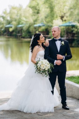 Beyaz elbiseli siyah saçlı bir gelin ve düğün günlerinde parkta papyonlu mavi elbiseli bir damat.