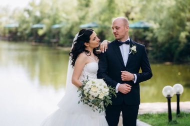 Beyaz elbiseli siyah saçlı bir gelin ve düğün günlerinde parkta papyonlu mavi elbiseli bir damat.