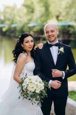 Beyaz elbiseli siyah saçlı bir gelin ve düğün günlerinde parkta papyonlu mavi elbiseli bir damat.