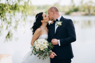 Beyaz elbiseli siyah saçlı bir gelin ve düğün günlerinde parkta papyonlu mavi elbiseli bir damat.