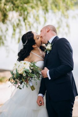 Beyaz elbiseli siyah saçlı bir gelin ve düğün günlerinde parkta papyonlu mavi elbiseli bir damat.