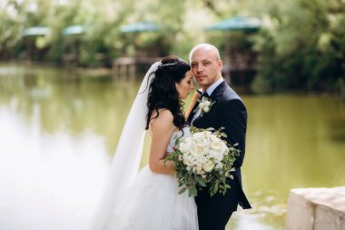 Beyaz elbiseli siyah saçlı bir gelin ve düğün günlerinde parkta papyonlu mavi elbiseli bir damat.